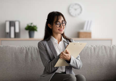 Focused arab female psychologist sitting on couch and writing in clipboard, offering professional counseling servicesの写真素材