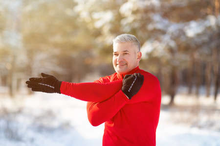 Happy mature male stretching his arm muscles, doing warmup exercises before jogging in snowy winter...