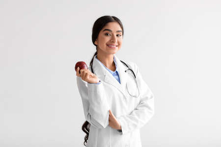 Happy pretty young indian woman doctor dentist in white coat with stethoscope hold fresh red appleの写真素材