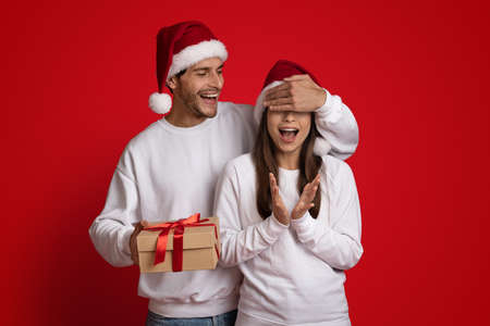 Cheerful Young Man In Santa Hat Making Christmas Surprise To Excited Girlfriendの写真素材