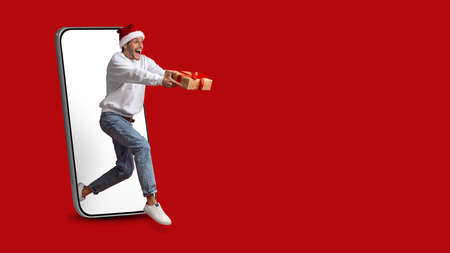 Christmas Delivery. Excited Guy With Gift Box Jumping Out Of Big Smartphone