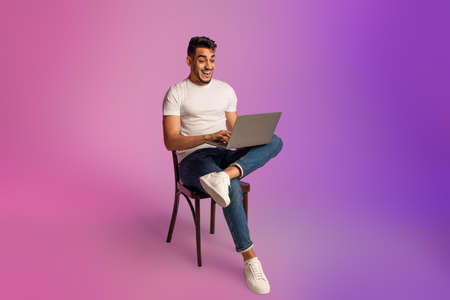 Shocked young Arab guy looking at laptop screen, sitting on chair, excited about online win or success in neon lightの写真素材