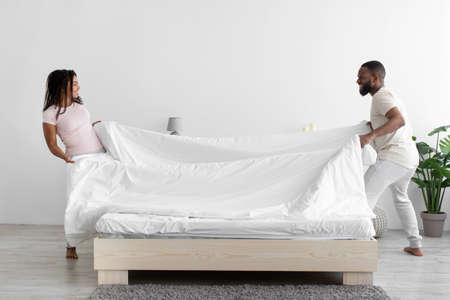 Cheerful millennial african american husband and wife make white bed with blanket in bedroom interiorの写真素材