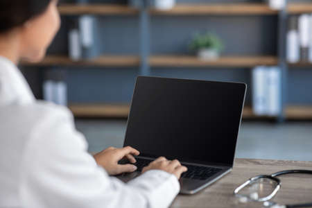 Young indian woman doctor in white coat typing on computer with empty screen in office interiorの写真素材