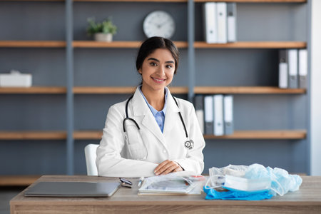 Smiling pretty young hindu woman doctor in white coat with stethoscope sits at table with many protective maskの写真素材