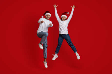 Cheerful Spouses Wearing Santa Hats Jumping In Air And Pointing At Cameraの写真素材