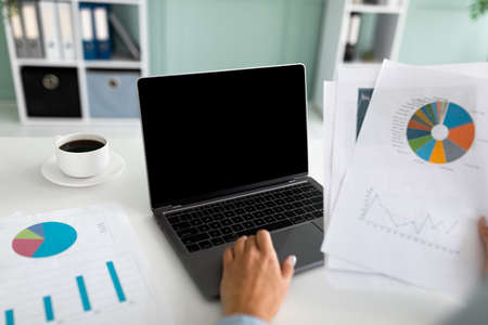 Female financial manager working on laptop computer with blank screen and doing paperwork, sitting...