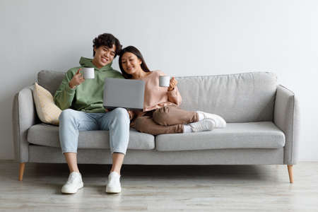 Happy Asian couple sitting on couch with laptop and coffee, browsing internet together at home, free spaceの写真素材
