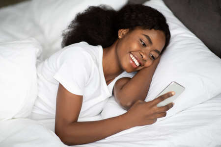 Positive young black lady laying in bed with smartphoneの写真素材