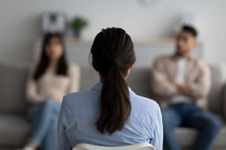 Female psychologist sitting with her back to camera, consulting young arab couple at office,...