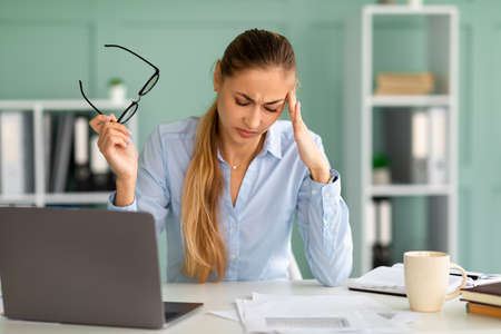 Stressed young businesswoman suffering from headache, sitting at workplace and touching temples,...