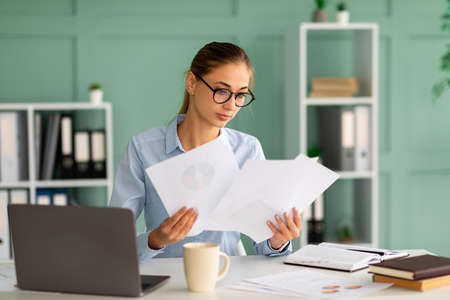 Concentrated businesswoman working with reports and documents, sitting with laptop at workplace in...