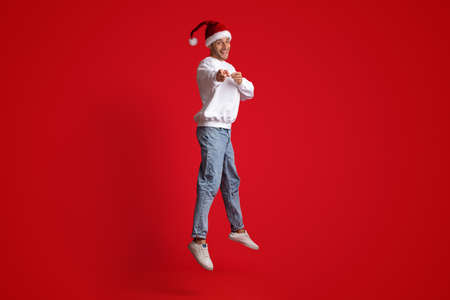 Cheerful Man Wearing Santa Hat Jumping In Air And Pointing At Cameraの写真素材