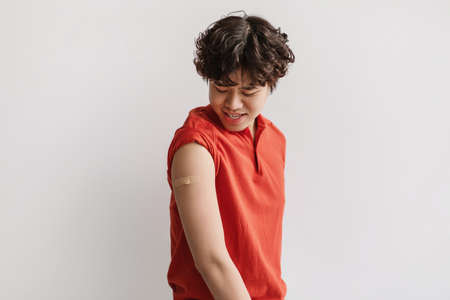 Cheerful vaccinated Asian man with plaster bandage on his shoulder after covid-19 vaccine injection...