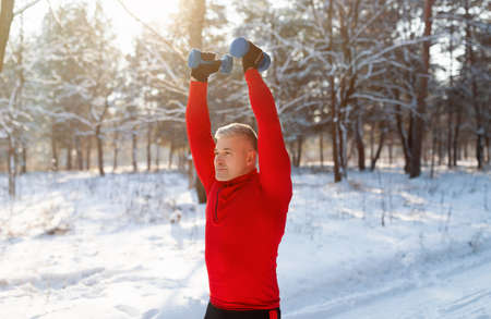 Outdoor winter sports. Athletic senior man working out with dumbbells, training arm muscles at snowy forestの写真素材