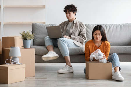 Frustrated young Asian woman unpacking carton box alone while her lazy boyfriend sitting on couch...