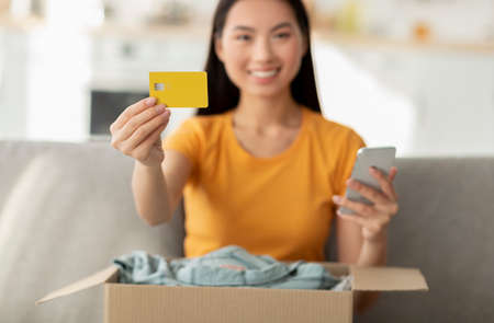 Online shopping and delivery service. Happy korean lady holding credit card and phone, unpacking box...
