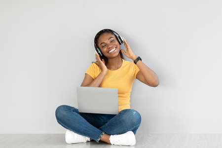 Young black woman using laptop computer, wearing wireless headphones, sitting on floor against grey...