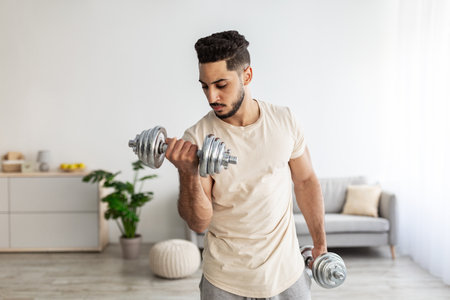 Home training concept. Handsome young Arab man exercising with dumbbells in living roomの写真素材