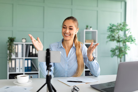 Remote learning concept. Happy teacher giving online lesson, using smartphone and laptop, sitting at workplace in officeの写真素材