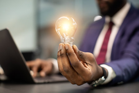 Black businessman holding illuminated light bulb, sitting in officeの写真素材