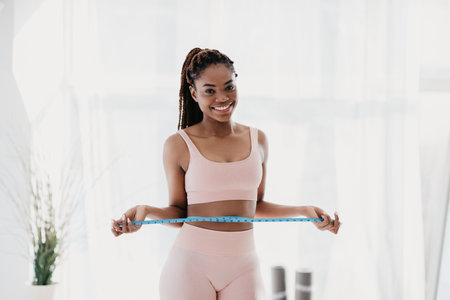 Young African American lady in sportswear measuring waist with tape, feeling happy about losing weight at homeの写真素材