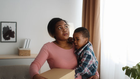 Black mother holding child and cardboard box walking in new flatの写真素材