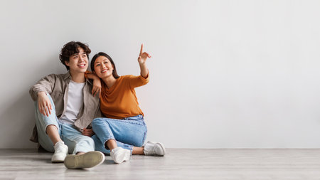 Carefree millennial Asian man and woman sitting on floor near white studio wall and pointing at copy...