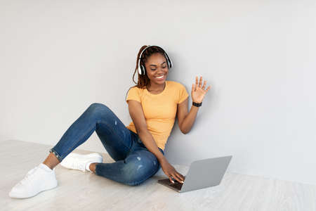 Young black lady using laptop pc for online communication, sitting on floor, waving at webcam...