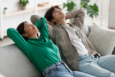 Peaceful young Asian couple relaxing on sofa with their hands behind heads in living roomの写真素材