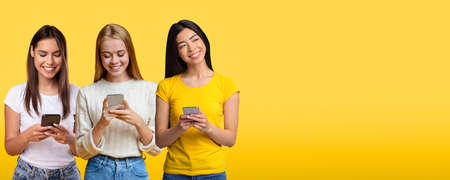 Three Young Women Using Smartphones While Standing Isolated Over Yellow Studio Background