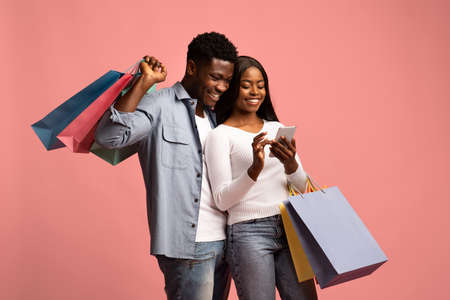 Happy black man and woman holding shopping bags, using smartphoneの写真素材
