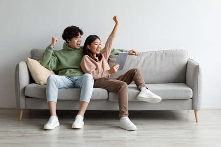 Excited Asian couple sitting on couch with tablet pc, celebrating online win, great deal or business success at homeの写真素材
