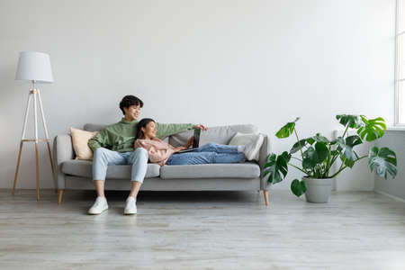 Cheery young Asian couple sitting on sofa, using laptop computer, communicating remotely, having video call at homeの写真素材