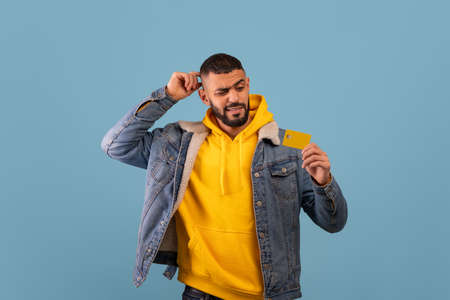 Young doubtful arab man holding credit card and thinking what to buy and touching head, blue...