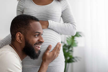 Glad happy millennial african american man listens to belly of pregnant wife in white bedroom interior, close upの写真素材