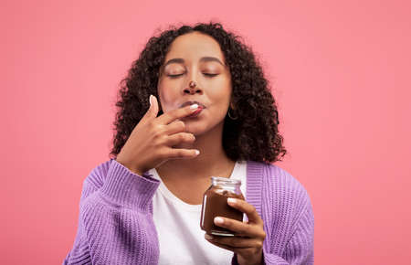 Black woman eating chocolate paste from jar, licking fingers, having dirty nose, enjoying dessert on...