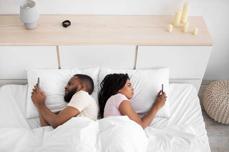 Sad young black guy and lady lie back to back, looks at smartphones on bed in bedroom interiorの写真素材