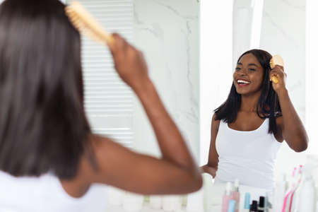 Beautiful African American Woman Combing Her Hair With Bamboo Brush In Bathroomの写真素材
