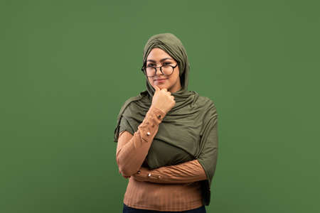 Pensive arab lady in glasses touching chin and thinking about question, standing over dark green...