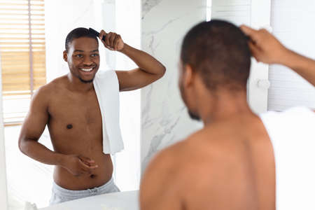 Handsome Shirtless Black Man Combing Hair And Looking In Mirror In Bathroomの写真素材