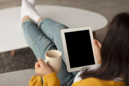 Millennial arab woman using digital tablet with blank screen and holding cup of tea, sitting on sofa at home, mockupの写真素材