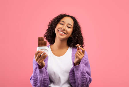 Cheat meal. Hungry African American lady holding chocolate bar, eating junk food over pink studio...
