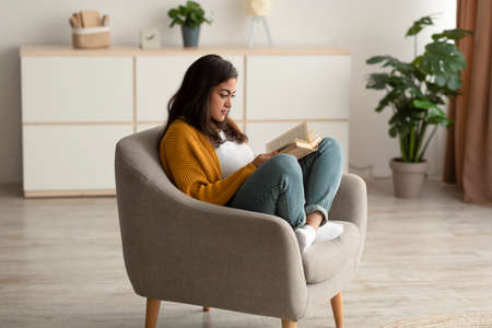 Focused arab lady sitting in armchair, reading book in living room, enjoying lazy morning with interesting storyの写真素材