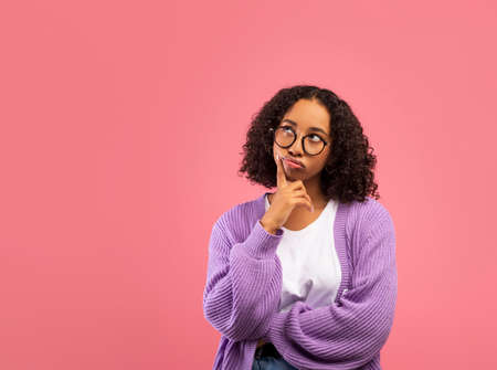 Portrait of pensive black woman touching chin and looking aside at copy space on pink studio...