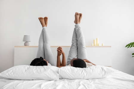 Glad happy young african american husband and wife in domestic clothes are lying on bed, holding hands with raised legsの写真素材