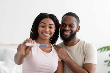 Satisfied attractive millennial black woman and guy show positive pregnancy test at camera in...