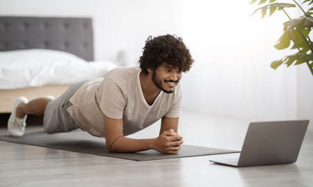 Happy athletic hindu man planking at home, using computer