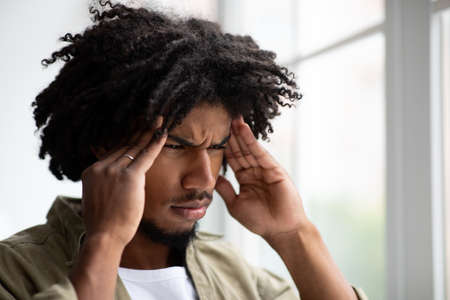Upset Young Black Guy Suffering From Headache While Standing Near Big Window