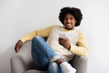 Nice App. Cheerful Black Millennial Guy Relaxing In Chair With Smartphone